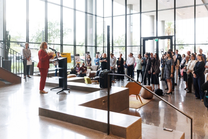 Woman speaking at a lectern to a crowd gathered in a lobby of a building.